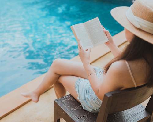 A woman is sitting by a pool with a tablet in her lap and a cup of coffee in front of her.