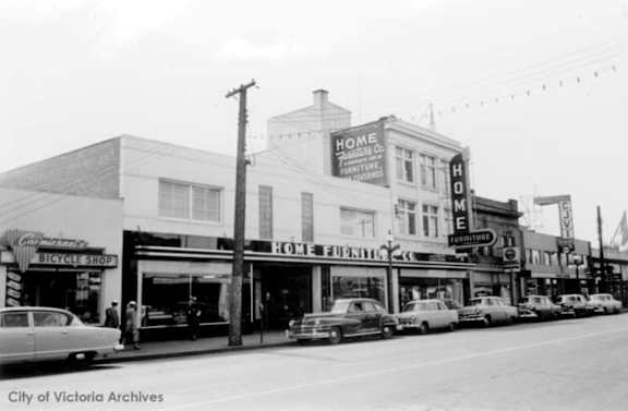 A black and white photo of a street with a Home Furnishings store and a Bicycle Shop.