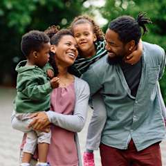 A family of four is standing outside with a car in the background.