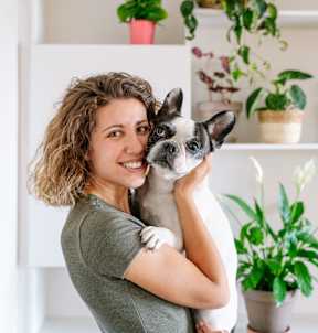 A woman holding a dog in front of a shelf with potted plants.