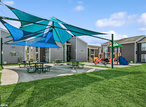 A playground with a blue and green umbrella and picnic tables.