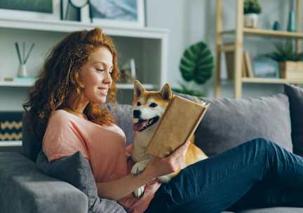 A woman is reading a book while sitting on a couch with her dog.