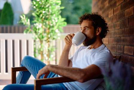 A man with curly hair is sitting on a chair, wearing a white t-shirt and blue jeans, holding a white mug.