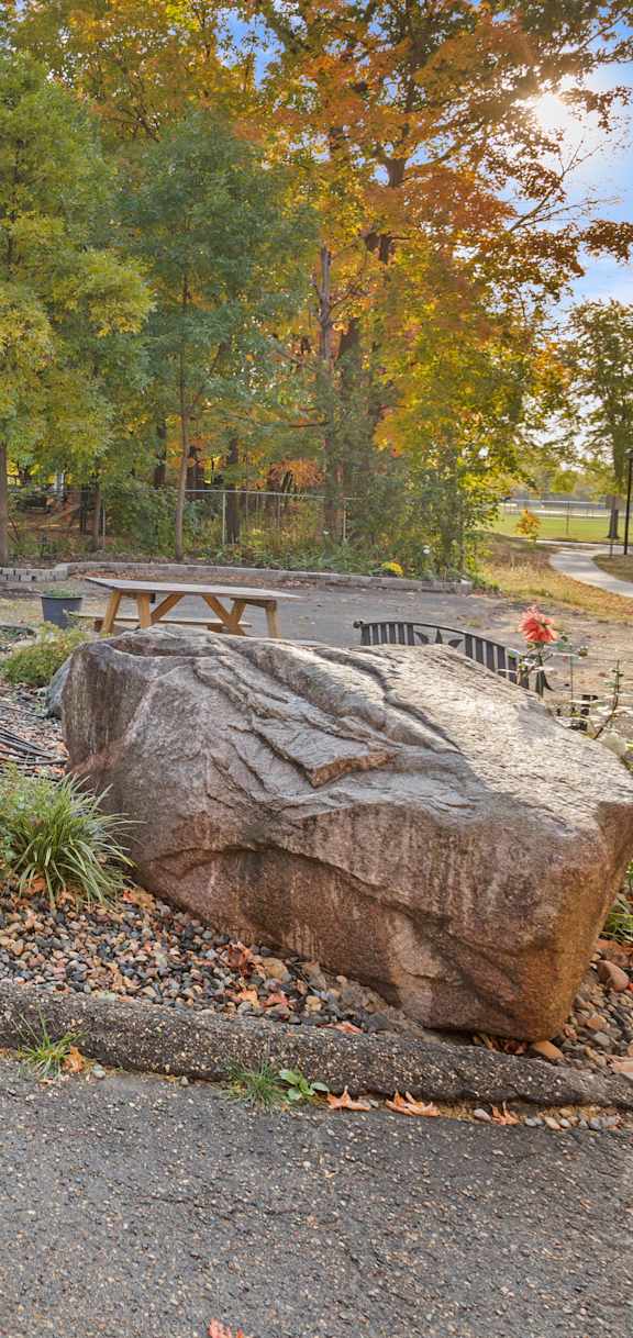 A large rock sits in the middle of a gravel area with a grassy area and trees in the background.
