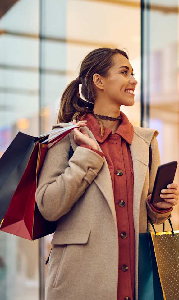 A woman is holding shopping bags and a phone while standing outside a store.