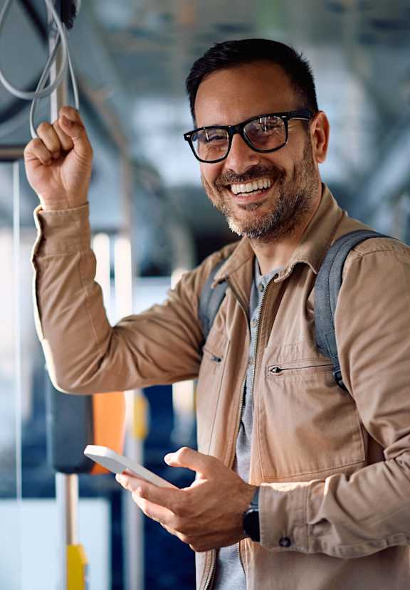 A man with glasses and a beard is smiling while holding onto a handle and looking at his phone.