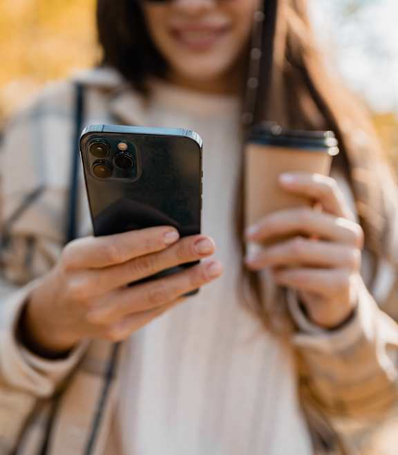A person in a beige jacket is holding a smartphone and a coffee cup.