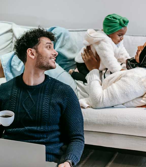A man is sitting on a couch with a laptop, holding a cup, and a woman is lying on the couch with a baby on her back.