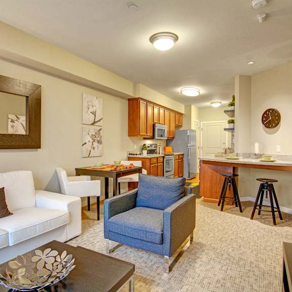 The spacious living room with a couch, plush chairs, coffee table, overhead lighting with a ceiling fan, decorations, a TV, and view of the kitchen with a bar with two barstools at the Artiste Apartment Homes.