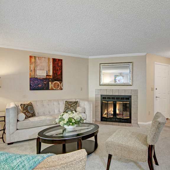The spacious, white living room with a white couch, throw pillows, large windows, a ceiling fan, wall art, plush white chairs, coffee table with white flowers, a fireplace, decorations, large windows, and a mirror at the Ridge and Shores Apartment Homes. 