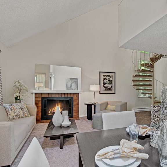 A living room with a cozy fireplace, white couch, gray chair, coffee table with white vases, lamps, wall art, decorations, a dining room with a dining table, chairs, and four places set, with a view of the kitchen and spiral staircase at the Cove Apartment Homes. 
