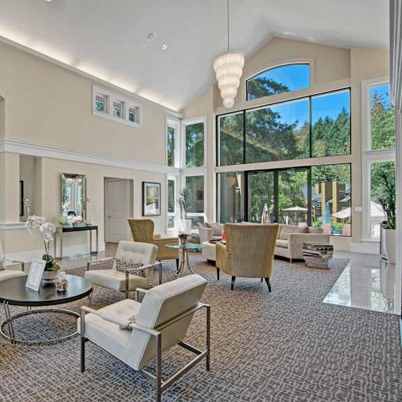 The resident lounge with white tables and chairs, large windows showing the sunny outdoors, coffee tables, and a TV at the Cove Apartment Homes. 