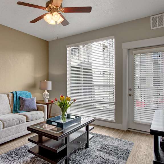 A living room with a tan couch with gray pillows and a blue throw blanket, a ceiling fan, glass doors to the balcony, and a coffee table at Velo on the Boulevard Apartment Homes. 