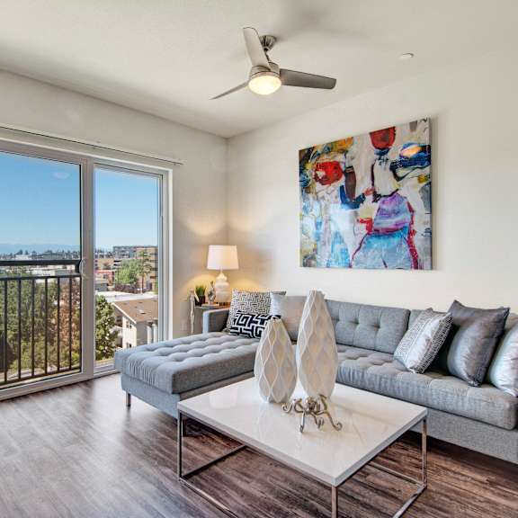 The spacious, bright living room with a couch, throw pillows, large windows, a ceiling fan, wall art, plush chairs, coffee table, decorations, a ceiling fan and lighting, and glass doors to the balcony at Vive Apartment Homes. 