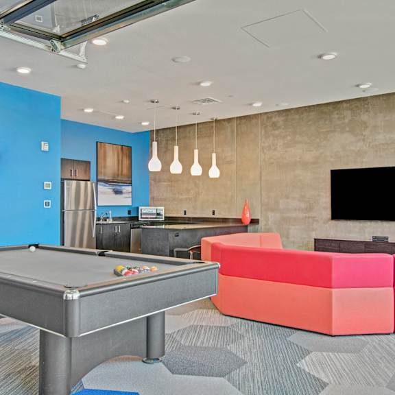 A wonderful resident lounge with tables, chairs, TVs, a pink couch, a pool table, a kitchenette, overhead lighting, and a bright blue accent wall at Vive Apartment Homes. 