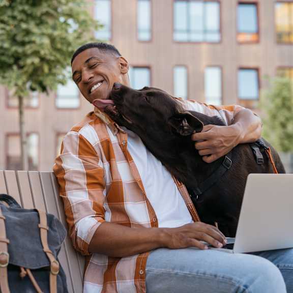 A man is sitting on a bench with a laptop and a dog is hugging him.