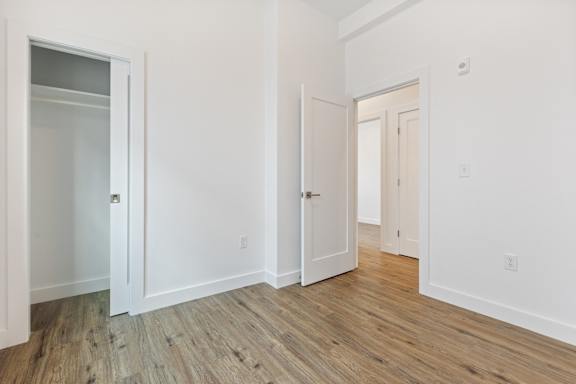 A room with white walls and wood flooring with a door on the left and a closet on the right.