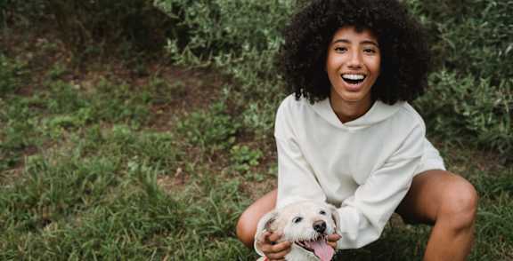 A woman in a white hoodie is sitting on the grass with a white dog wearing a red harness.