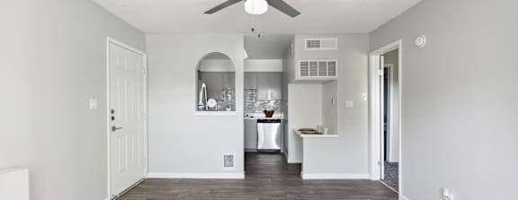 A room with a ceiling fan and a kitchen area in the background.