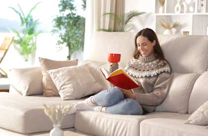A woman is sitting on a beige couch reading a red book.