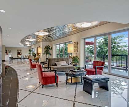 The amazing resident lounge with tan couches, decorative potted plants, decorations, large windows, unique overhead lighting, sliding glass doors to the balcony, red plush chairs, and coffee tables at the Artiste Apartment Homes. 