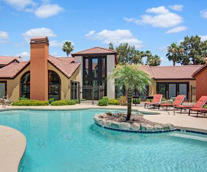 The turquoise outdoor pool with the main office and lounge in the background, pool chairs, tables, and chairs with the apartments in the background at Motif Apartments.