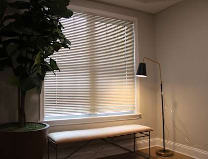 A potted plant sits next to a window with blinds, a bench is placed in front of the window, and a floor lamp stands to the side.