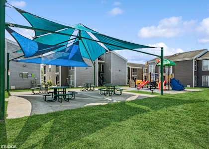 A playground with a blue and green umbrella and picnic tables.