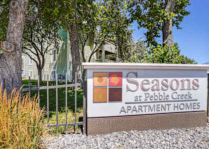 A sign for Seasons at Pebble Creek Apartment Homes is in front of a fence and trees.