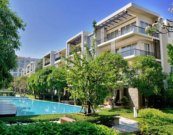 A swimming pool is surrounded by green trees and bushes in front of a building.
