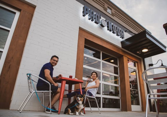 a man and woman sit at a table outside a restaurant with their dog