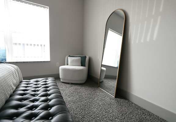 A large mirror sits on a grey carpeted floor next to a white chair and a bed.