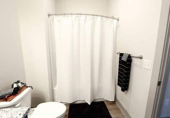 A white shower curtain in a bathroom with a black towel hanging on the wall.