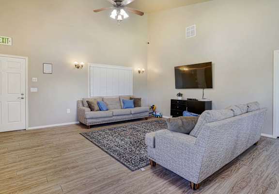 A living room with a grey couch, a rug, a ceiling fan, and a flat screen TV mounted on the wall.