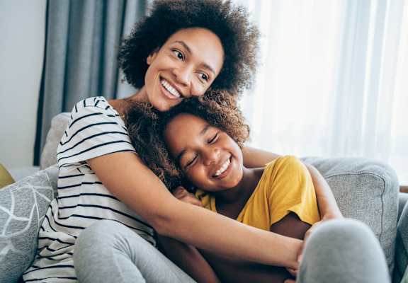 A woman with curly hair is hugging a child on a grey couch.