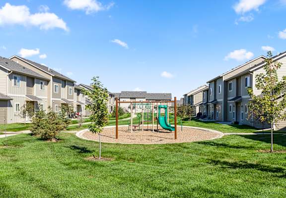 A playground with a green slide in the middle of a grassy area surrounded by houses.