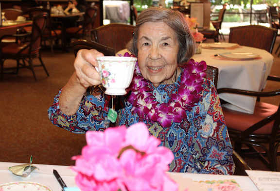 An elderly woman in a floral shirt is holding a cup and smiling.
