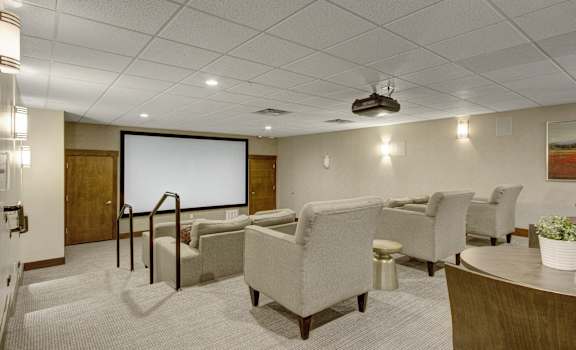 The entertainment room with a movie theater screen, plush white chairs, plush white couches, wall art, and coffee tables at Velo on the Boulevard Apartment Homes. 