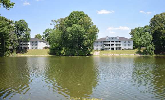 A large white building sits on the edge of a lake surrounded by trees.