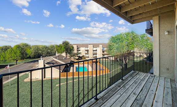 A balcony with a black railing and a wooden floor overlooks a green lawn and a building.