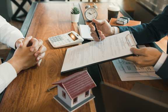 A person is signing a document with a house model and a calculator on the table.
