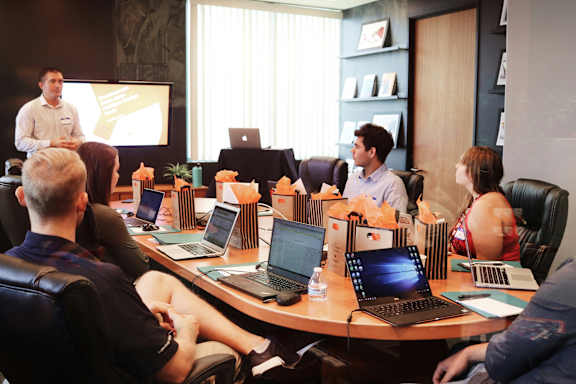 A group of people are sitting around a table with laptops and papers, listening to a man giving a presentation.