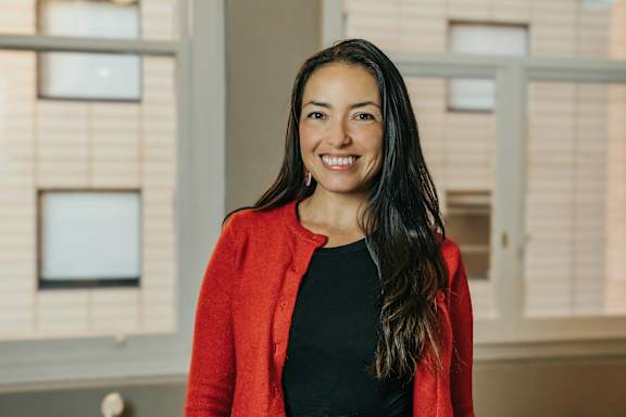 A woman with long dark hair wearing a red cardigan and black top standing in front of a window.