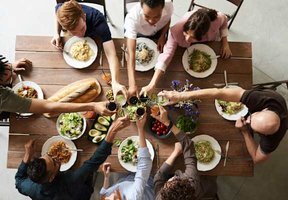 A group of people sitting around a table with plates of food, reaching out to each other.