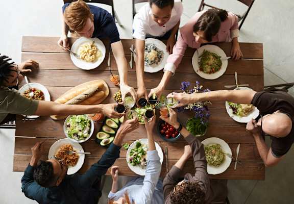 A group of people sitting around a table with plates of food, reaching out to each other.