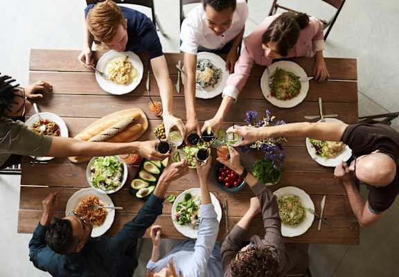 A group of people sitting around a table with plates of food, reaching out to each other.