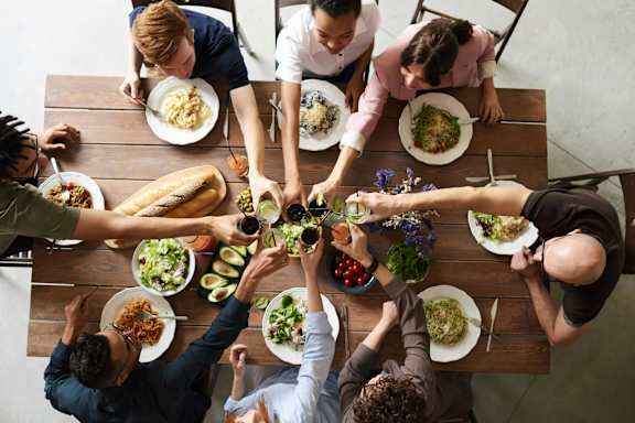A group of people sitting around a table with plates of food, reaching out to each other.