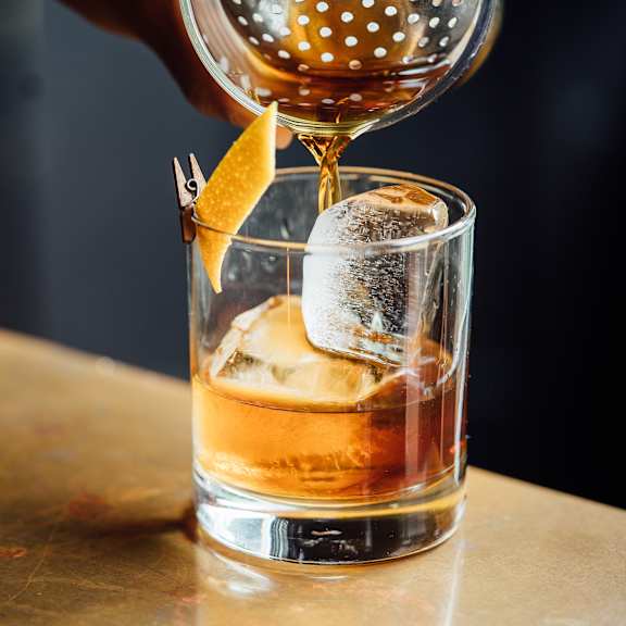 A glass of whiskey with ice and a lemon peel garnish is being filled with more whiskey from a decanter.