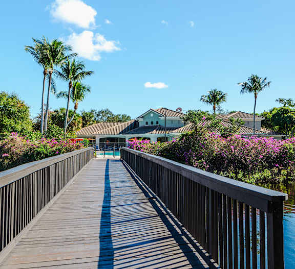 A wooden walkway leads to a building with a white roof and a green lawn with purple flowers.