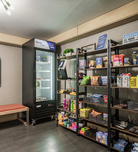A vending machine is in the middle of a room with a bench and a picture on the wall.
